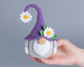 A crocheted gnome doll with a purple hat, white beard, and daisy details, held in a person's hand against a plain gray background.