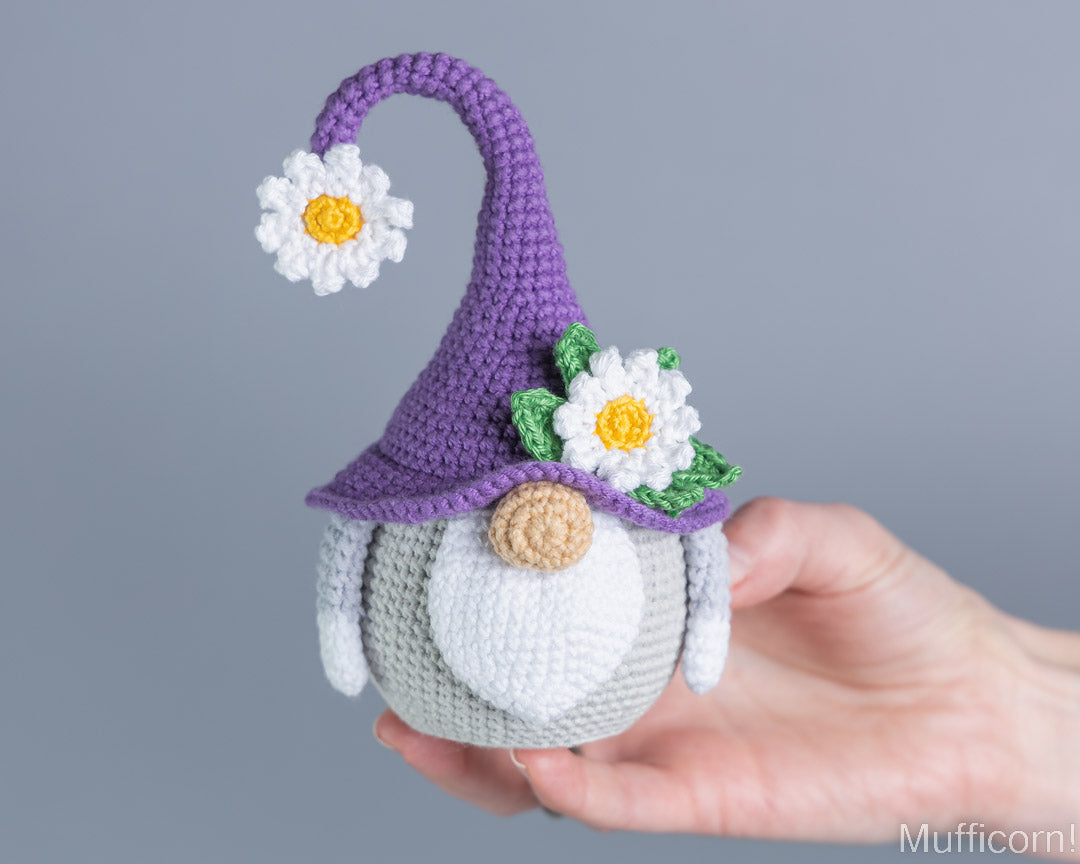 A crocheted gnome doll with a purple hat, white beard, and daisy details, held in a person's hand against a plain gray background.