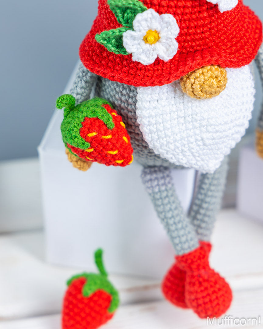A close-up of a crocheted gnome doll with a red hat adorned with a white flower, a white beard, gray legs, red shoes, and a small strawberry in one hand. Another strawberry lies on the white surface below.