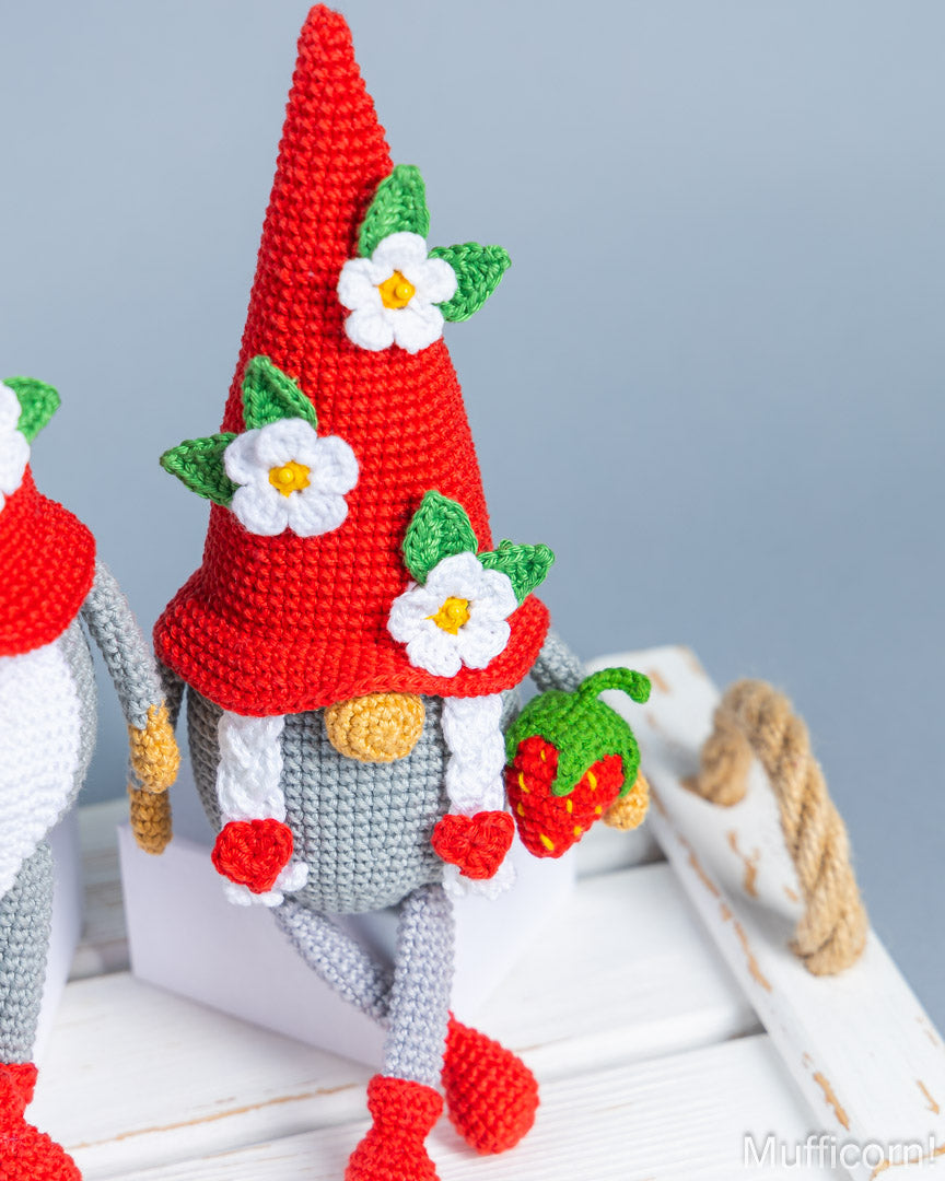 A crocheted gnome doll with a tall red hat decorated with white flowers and green leaves, gray body and legs, red boots, and a strawberry accessory, posed on a white wooden surface against a soft blue-gray background.
