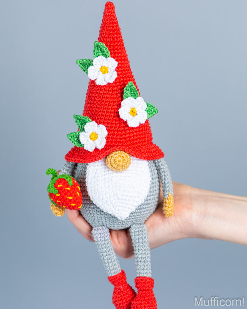 A crocheted gnome doll with a tall red hat, white flowers, green leaves, a white beard, gray body and legs, red shoes, and a small strawberry accessory, held in a person's hand against a plain gray background.
