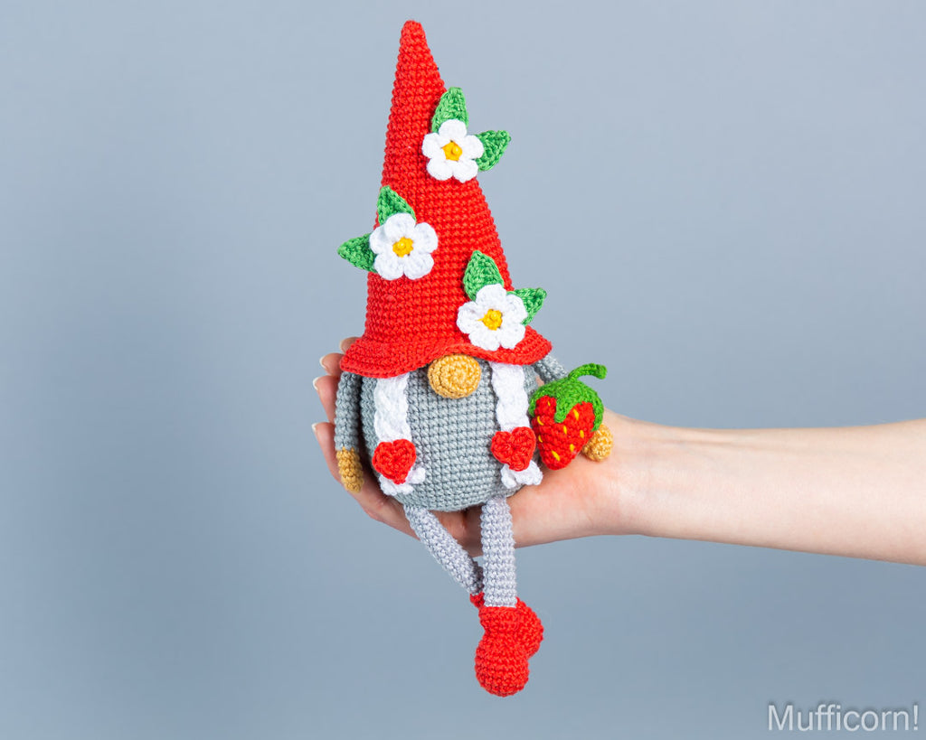 A crocheted gnome doll with a tall red hat, white flowers, green leaves, and red boots is being held in a person's hand against a plain light blue-gray background.