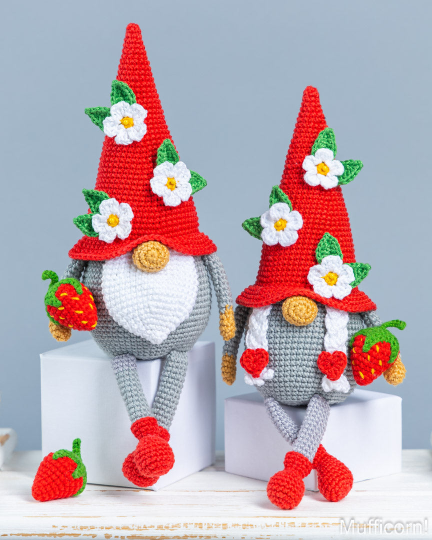 Two crocheted gnome figurines with red pointed hats decorated with white flowers and green leaves, gray bodies, white beards, and red-and-green strawberry accessories, displayed on white blocks against a light blue-gray background.