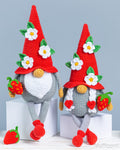 Two crocheted gnome dolls with red pointed hats, white flower accents, green leaves, gray bodies, and red-and-green strawberry accessories, posed on white blocks against a light blue-gray background.