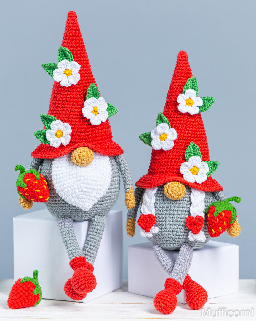 Two crocheted gnome dolls with red pointed hats, white flower accents, green leaves, gray bodies, and red-and-green strawberry accessories, posed on white blocks against a light blue-gray background.