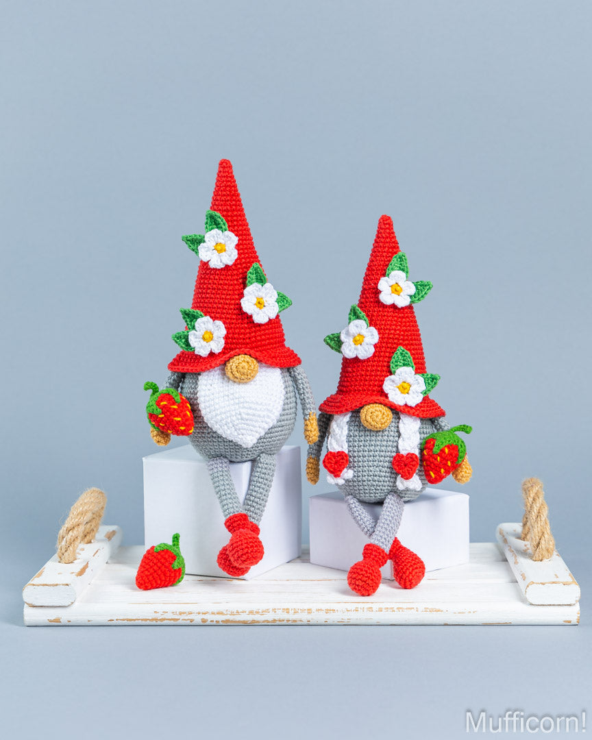 Two crocheted garden gnome figures with red pointed hats decorated with white flowers, gray bodies, and red-and-green strawberry accents, posed on a white rustic tray against a light blue-gray background.