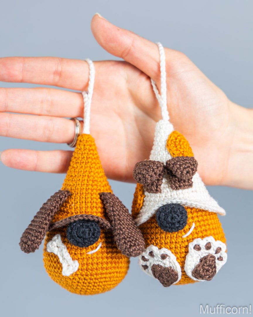 Two crocheted dog-shaped ornaments held in a person's hand against a plain light gray background. The ornaments are mustard yellow with brown and white details, including floppy ears, black eyes, a bone, and paw prints.