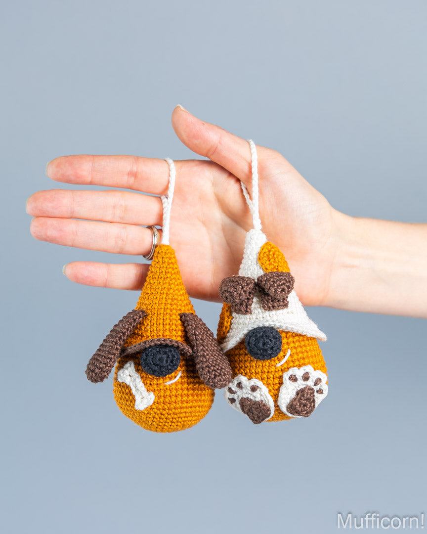 A hand holds two small crocheted dog-shaped ornaments against a plain light gray background. The ornaments are orange with white and brown details, including floppy ears, a black nose, and paw prints.