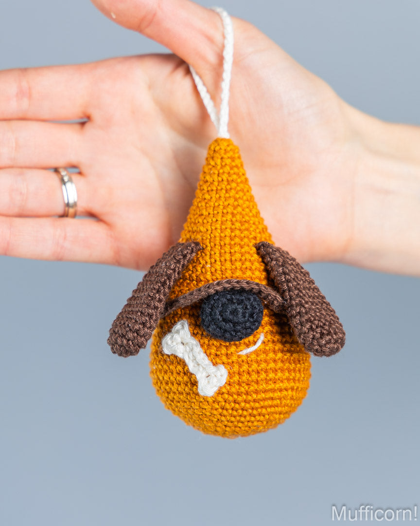 A crocheted dog-shaped ornament held in a person's hand against a plain gray background. The ornament is mustard yellow with brown floppy ears, a black stitched eye, a white bone detail, and a white hanging loop.