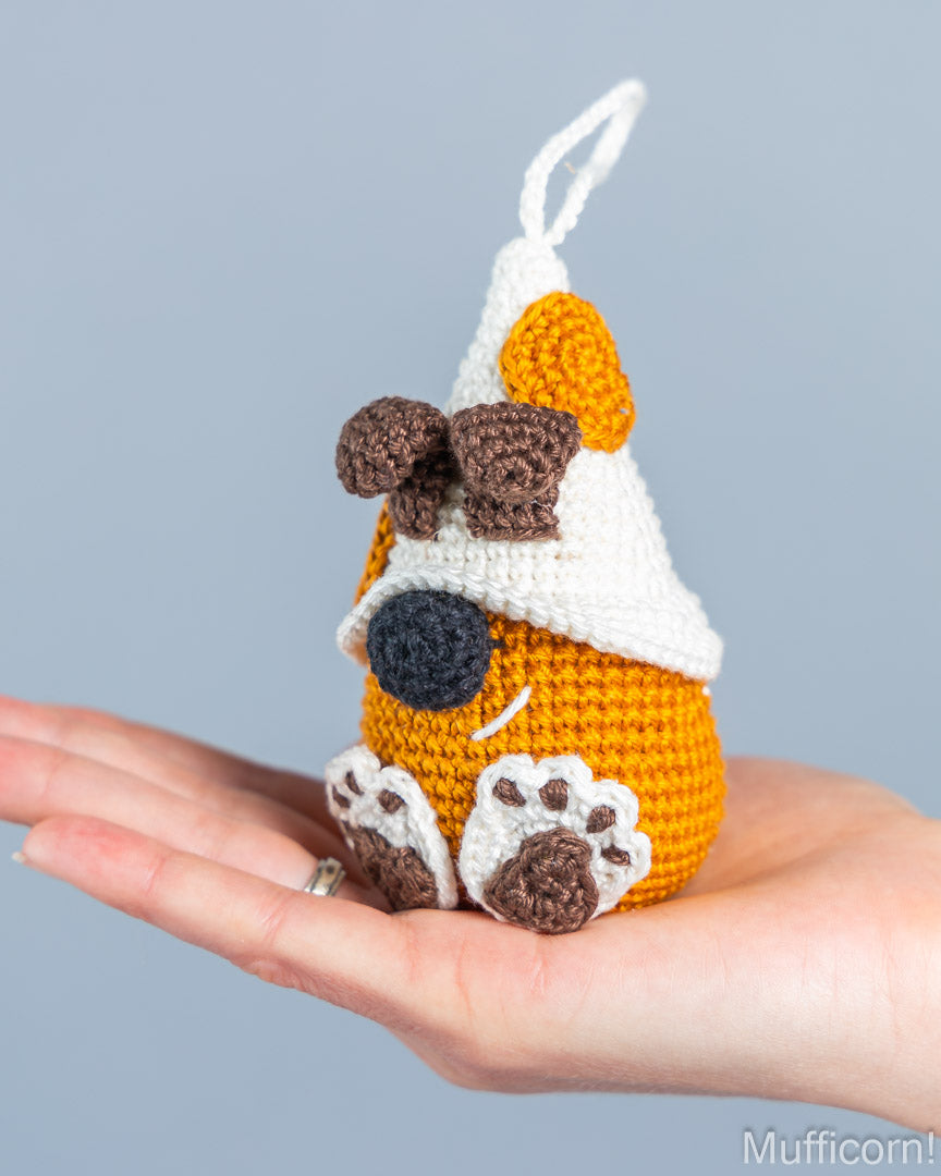 A small crocheted dog-shaped ornament or plush toy sits on a person's hand against a plain light gray background. The toy is primarily orange with a white hat, brown ears and paw pads, a dark gray nose, and white paw prints on the feet.