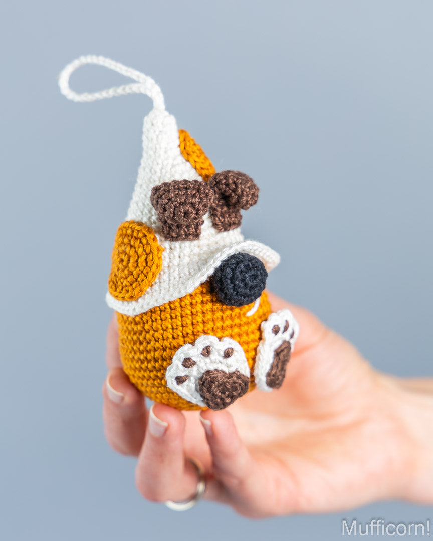 A hand holds a small crocheted dog-shaped ornament or charm against a soft light blue-gray background. The ornament is primarily mustard yellow and white, with brown and black details, and has a white hanging loop at the top.