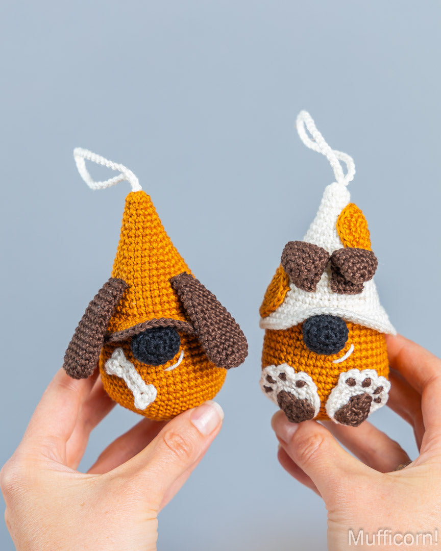 Two handmade crocheted dog ornaments held in hands against a light gray background. One ornament is orange with brown floppy ears and a white bone detail, while the other is orange and white with a brown bow, white paw pads, and white hanging loops.