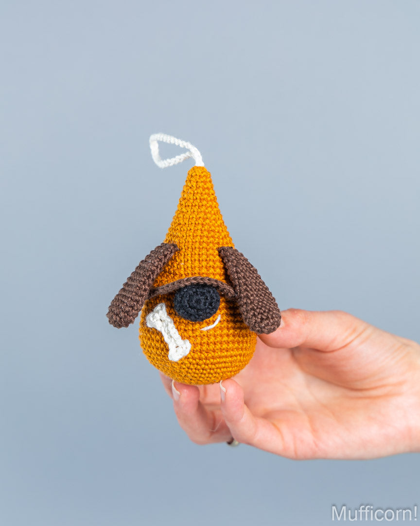 A hand holding a crocheted dog-shaped ornament or charm against a plain light gray background. The ornament is primarily mustard yellow with brown floppy ears, a black eye patch, a white bone detail, and a small white hanging loop at the top.