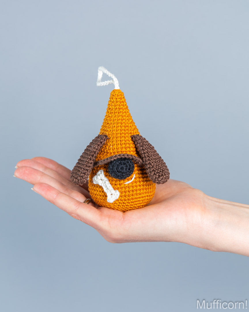 A small crocheted dog toy with floppy brown ears, a white bone, and a white triangular hat is held in a person's hand against a plain light gray background.