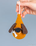 A hand holds a crocheted dog-shaped ornament against a plain light gray background. The ornament is mustard yellow with brown floppy ears, a black nose, and a white bone detail.