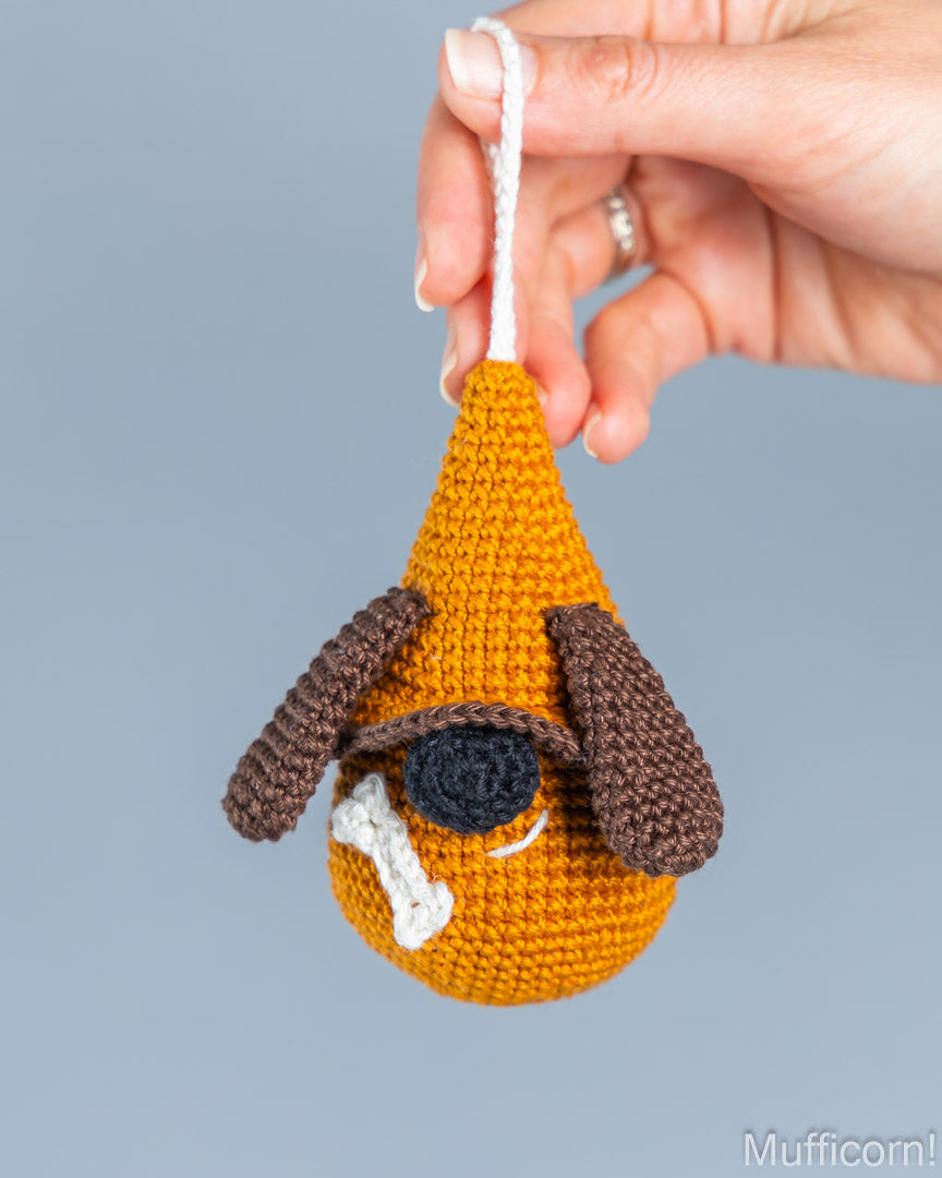 A hand holds a crocheted dog-shaped ornament against a plain light gray background. The ornament is mustard yellow with brown floppy ears, a black nose, and a white bone detail.