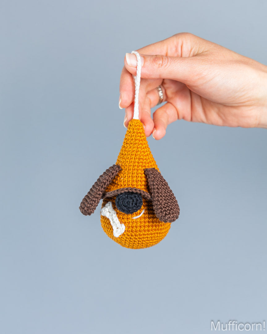 A hand holds a small crocheted dog-shaped ornament against a plain light blue-gray background. The ornament is mustard yellow with dark brown floppy ears, a black nose, and a white hanging loop.