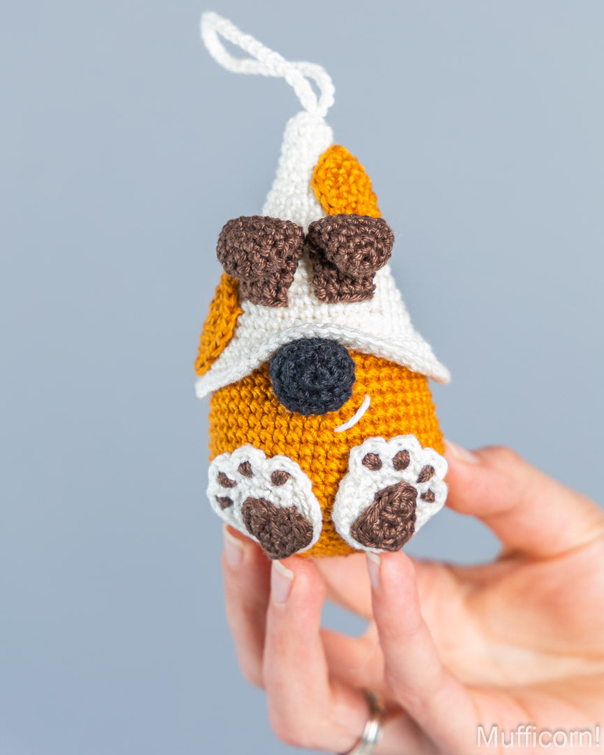 A hand holds a crocheted dog-shaped ornament or charm with a white hanging loop, featuring an orange body, white and brown pointed ears, a black nose, and white paw pads with brown centers, against a plain light gray background.