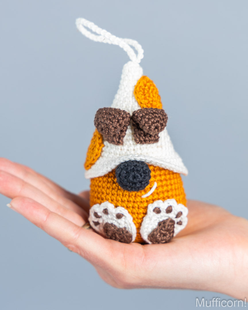A small crocheted dog-shaped ornament or plush toy is held in a person's hand against a soft light gray background. The toy is primarily orange with a white hat, brown ears and paw pads, a black nose, and a white hanging loop.