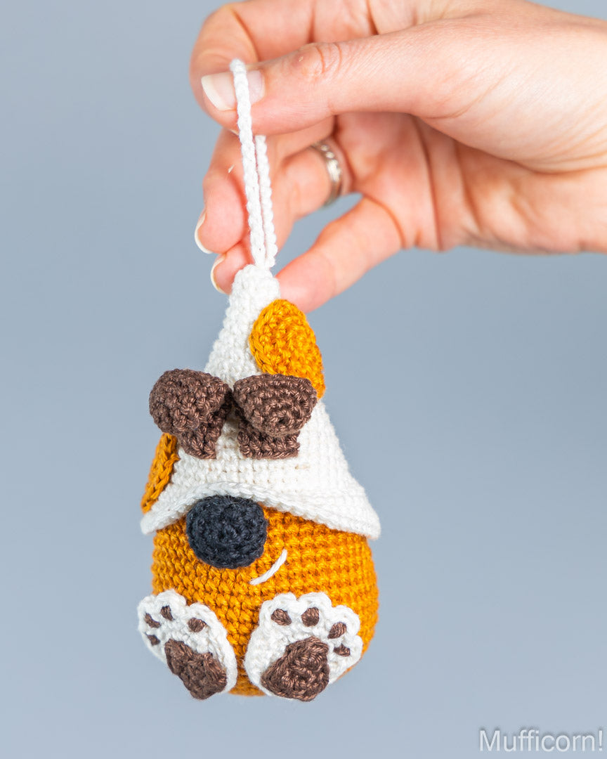 A hand holds a small crocheted dog-shaped ornament or charm against a plain light gray background. The ornament is primarily orange with white, brown, and black details, including a white hanging loop, white paws, and a black nose.