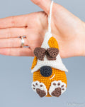 A hand holds a small crocheted dog-shaped ornament or charm against a plain light gray background. The dog has an orange body, white hat and paw pads, brown ears and paw prints, and a black nose.