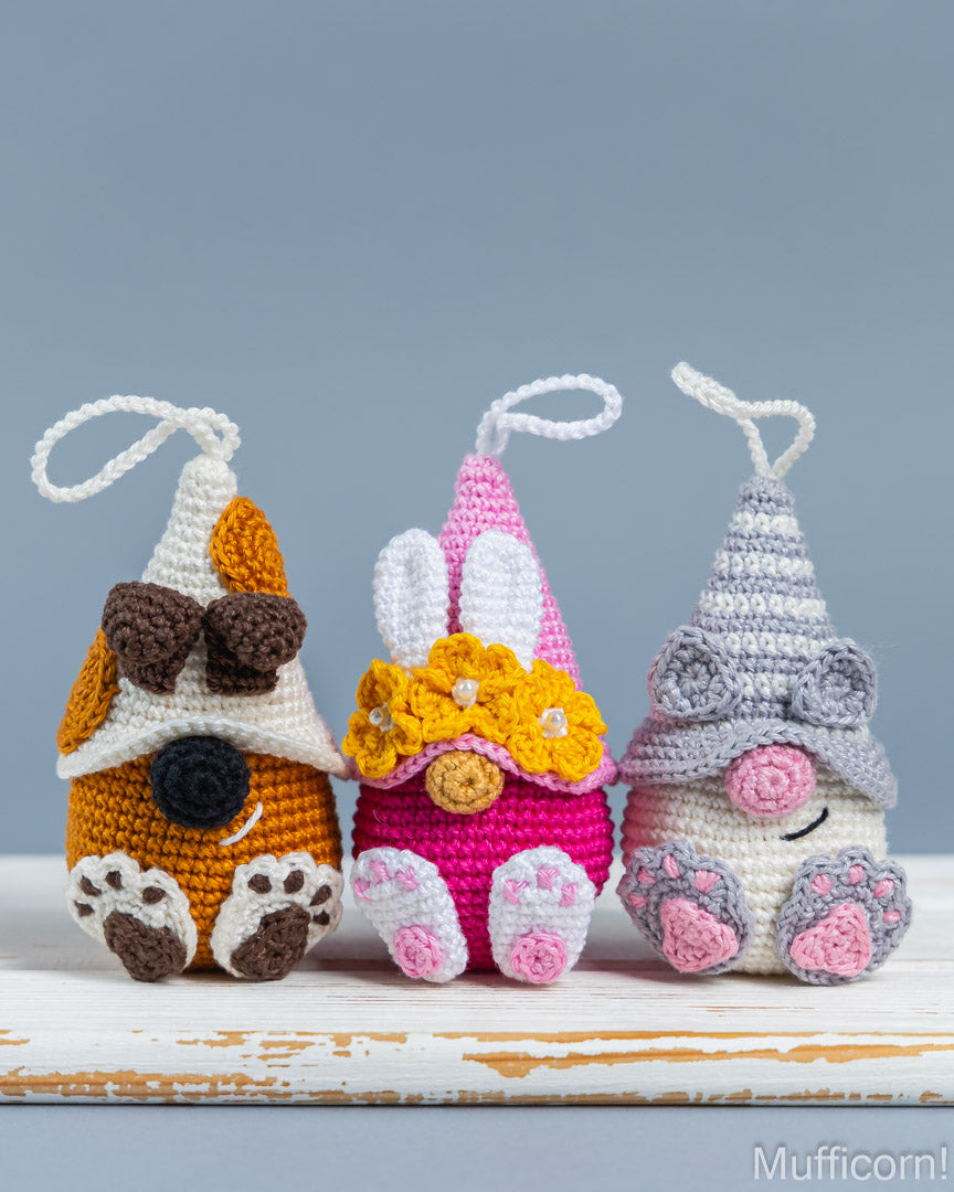Three handmade crocheted gnome ornaments in orange, pink, and gray, lined up on a white rustic wooden surface against a solid blue-gray background.