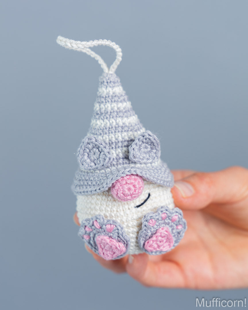 A small crocheted gnomelike ornament or plush charm with a pointed lavender hat, white body, pink nose, and pink paw pads, being held in a person's hand against a soft blue-gray background.