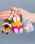 Three small crocheted animal-shaped pouches or charms are held in a person's hand against a plain gray background. The left one is orange and white with brown paw prints and a black circular detail; the middle one is pink, yellow, and white with bunny ears and flowers; the right one is gray, cream, and pink with cat-like features.
