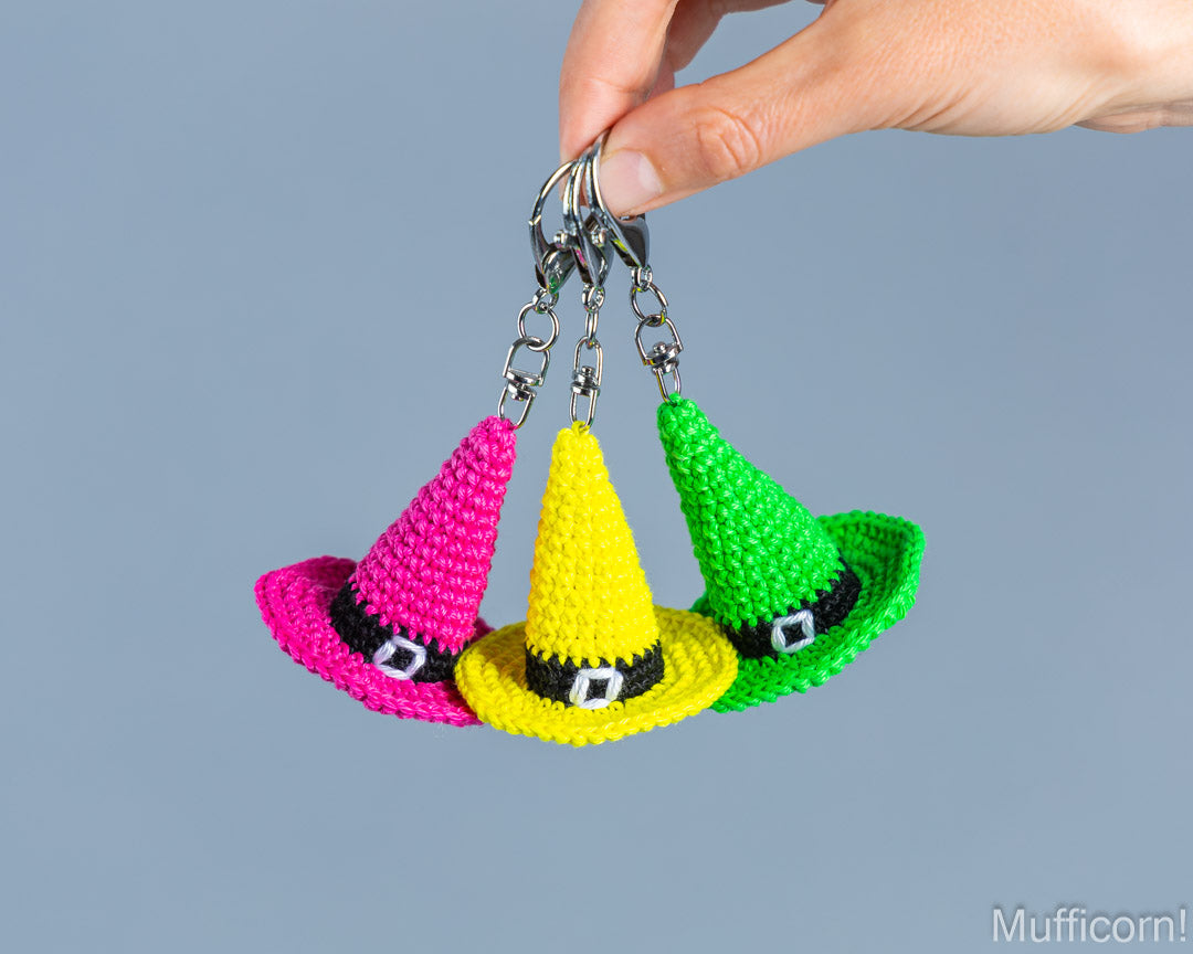 Three small crocheted witch hats keychains in pink, yellow, and green, held by a hand against a solid blue-gray background.