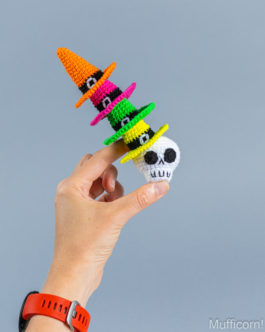 A hand holds a crocheted Halloween-themed finger toy featuring a white skull wearing a stack of colorful witch hats in orange, pink, green, yellow, and black, against a solid gray background.
