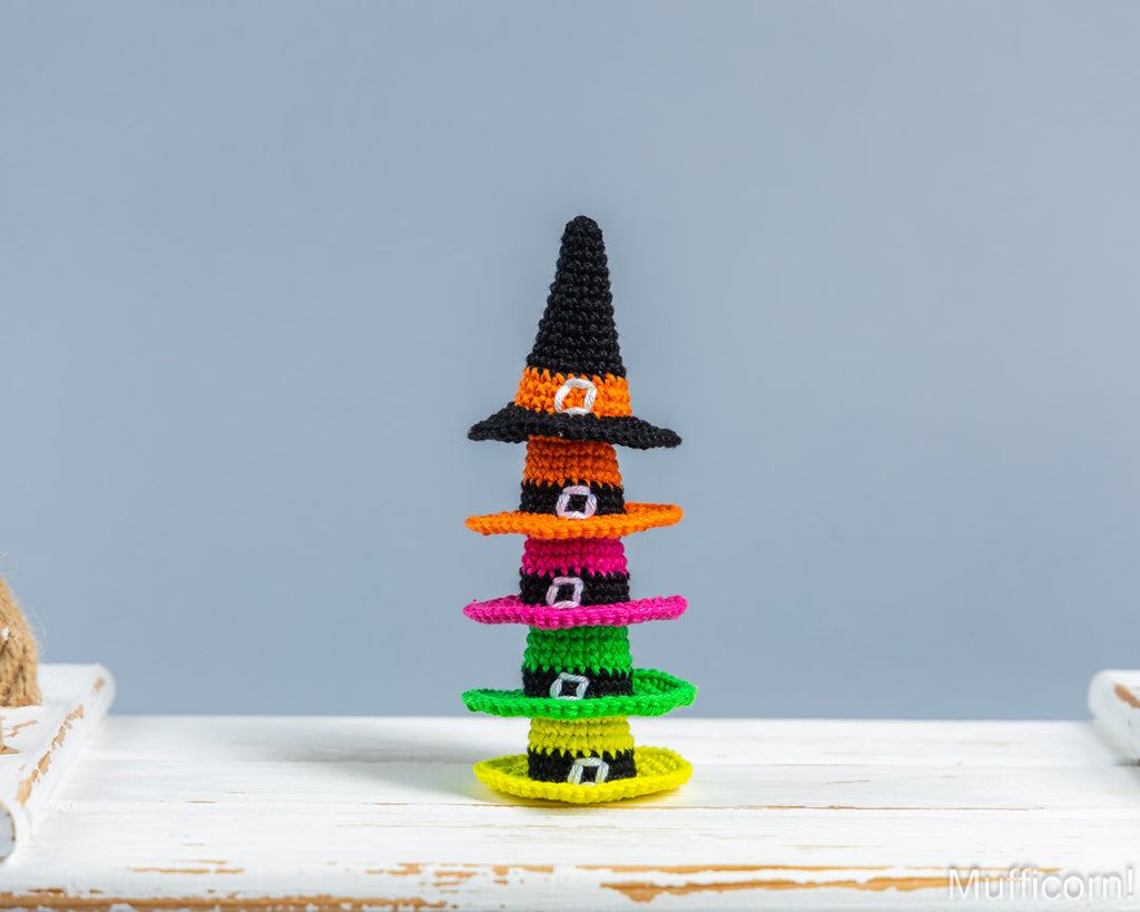 A small crocheted Halloween witch hat stack decoration made of layered beaded hats in black, orange, pink, green, and yellow, displayed on a white wooden surface against a light blue-gray background.