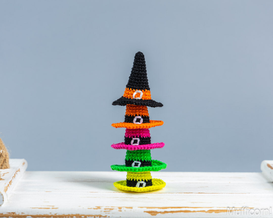 A small crocheted Halloween witch hat stack decoration made of layered beaded hats in black, orange, pink, green, and yellow, displayed on a white wooden surface against a light blue-gray background.