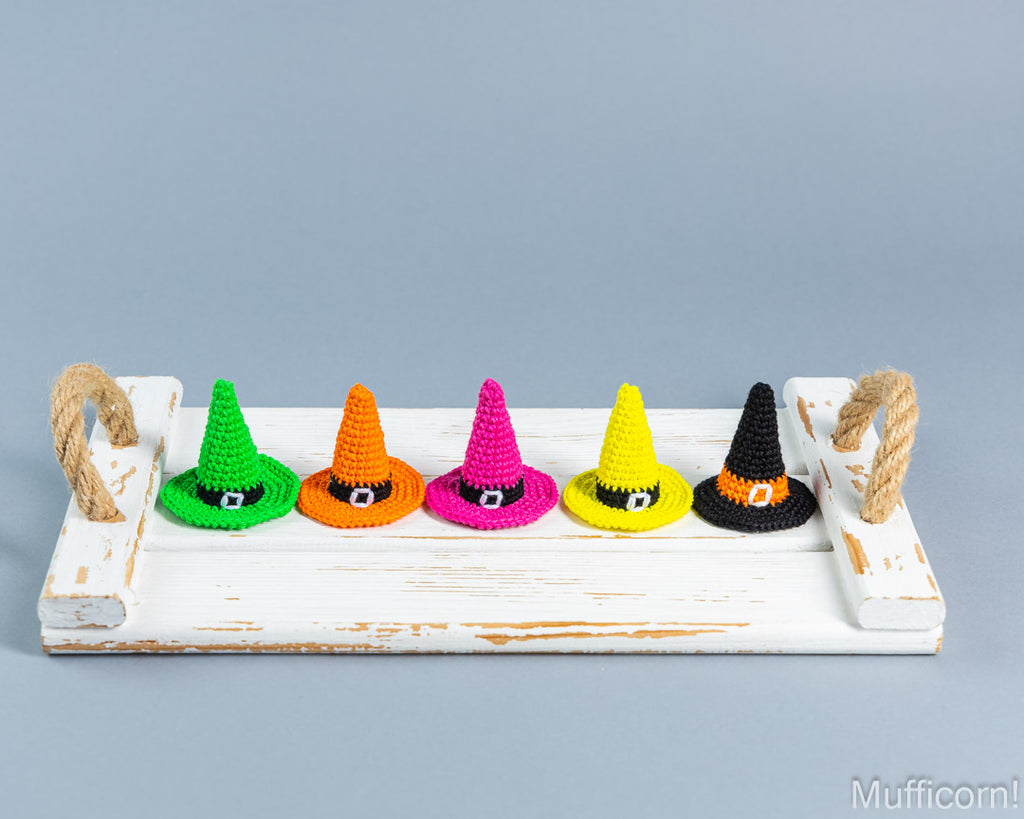 Five crocheted witch hats in green, orange, pink, yellow, and black are lined up on a distressed white wooden tray with rope handles against a solid gray background.