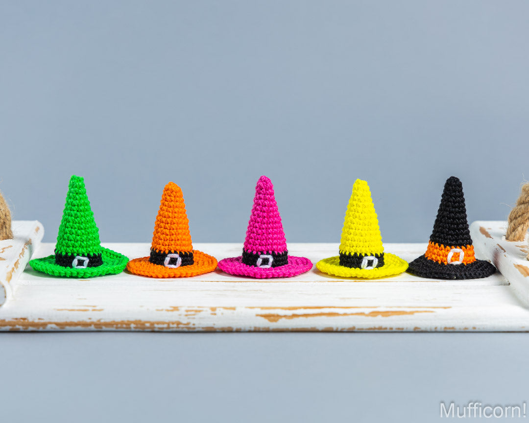 Five small crocheted witch hats in green, orange, pink, yellow, and black, arranged in a row on a white rustic board against a light blue-gray background.