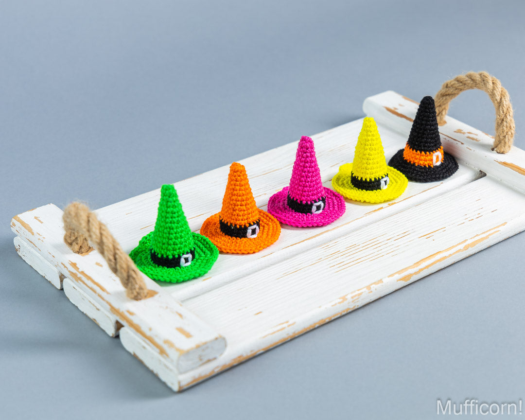 Five crocheted witch hats in green, orange, pink, and yellow, with a black and orange hat on the right, arranged on a distressed white wooden tray with rope handles against a gray background.