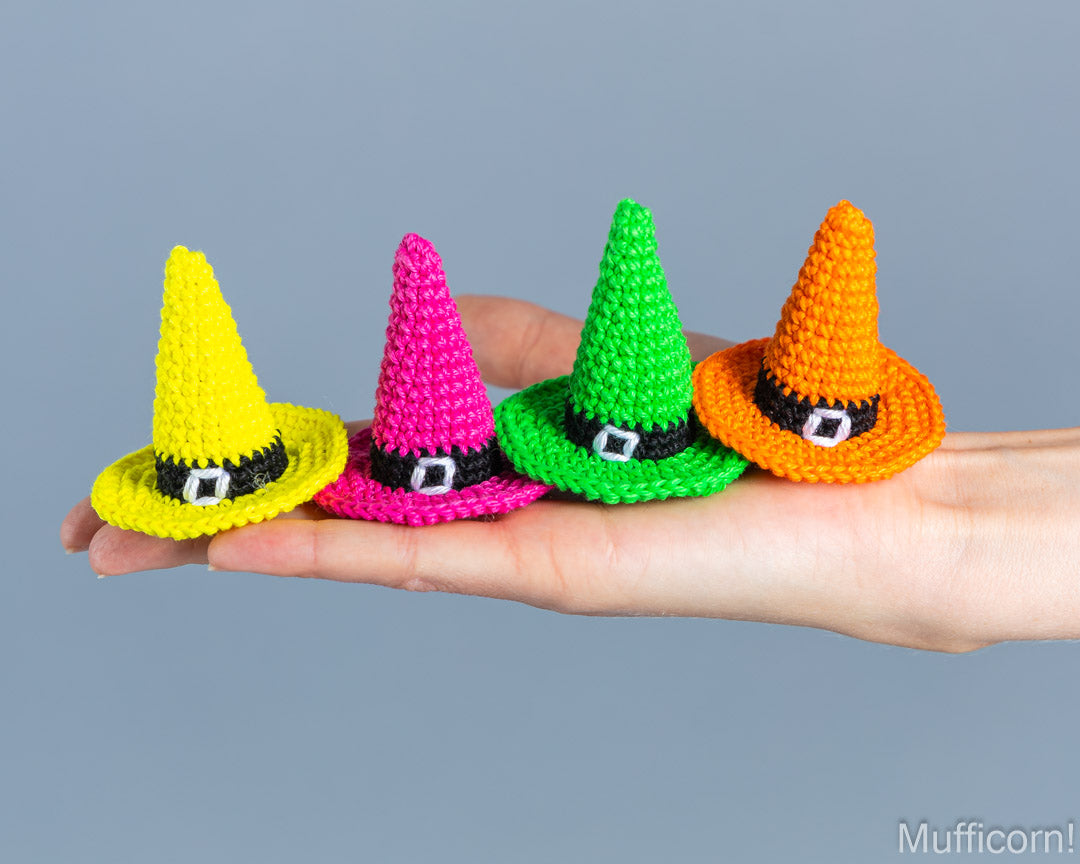Four small crocheted pointed hats in yellow, pink, green, and orange are arranged on an open hand against a plain blue-gray background.