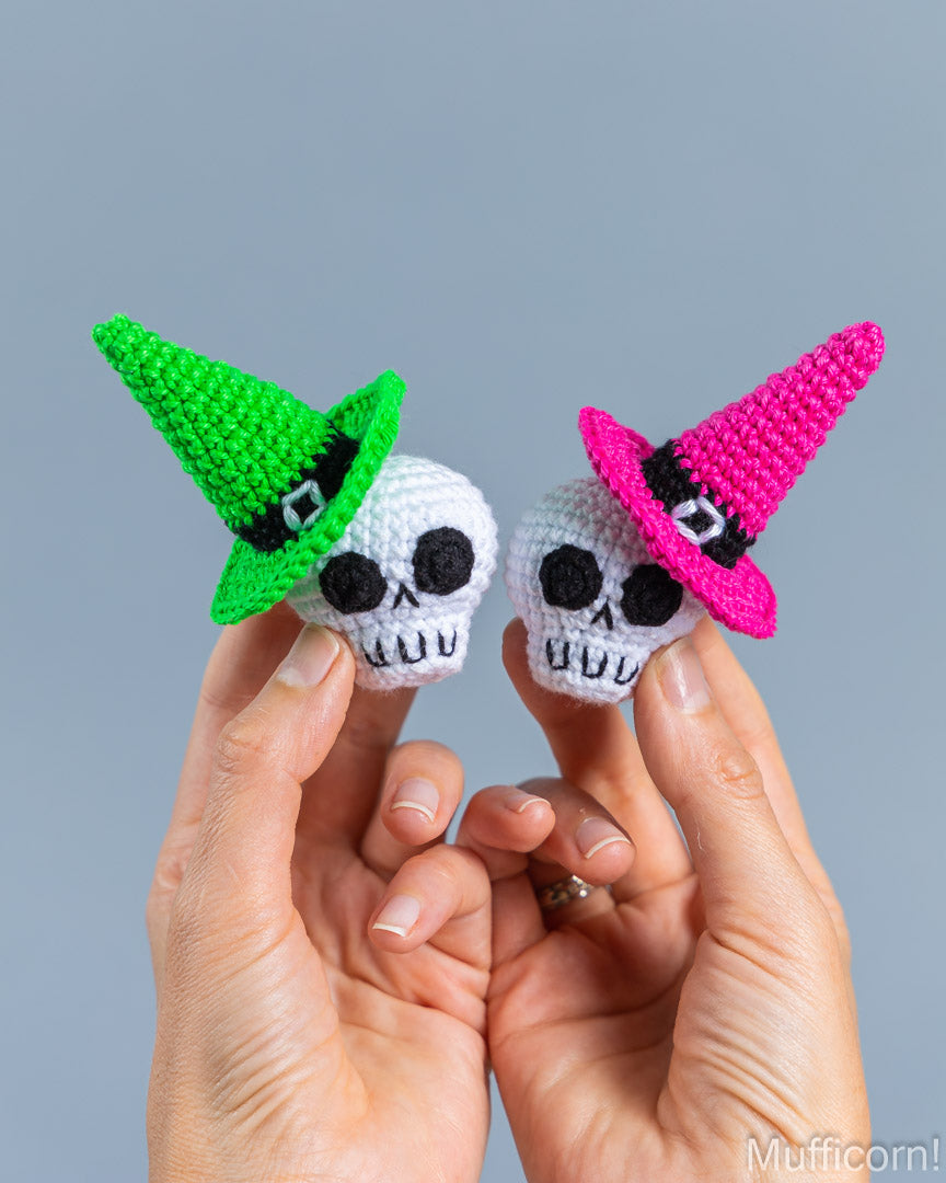 Two small crocheted skull figurines wearing pointed witch hats are held between two hands against a plain light gray background. One hat is bright green and the other is bright pink, and both skulls are white with black facial details.