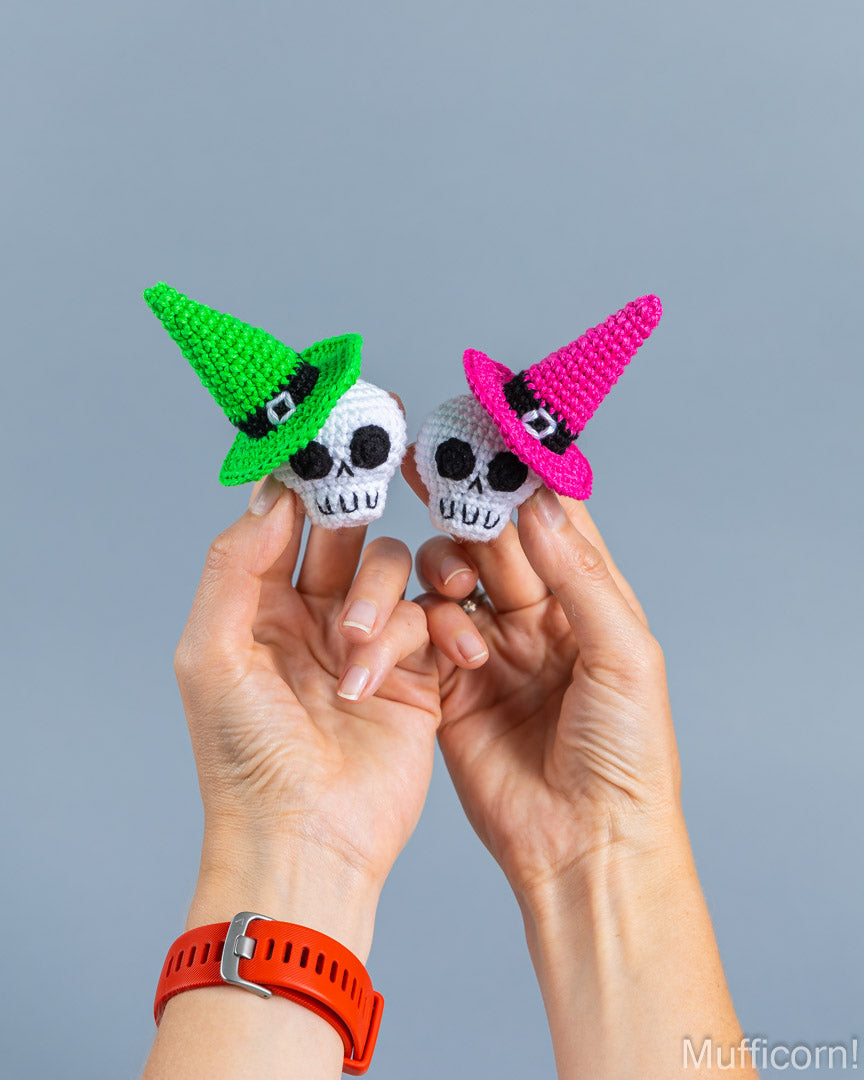 Two hands holding two small crocheted skull-shaped accessories with pointed witch hats, one in green and one in pink, against a plain gray background.