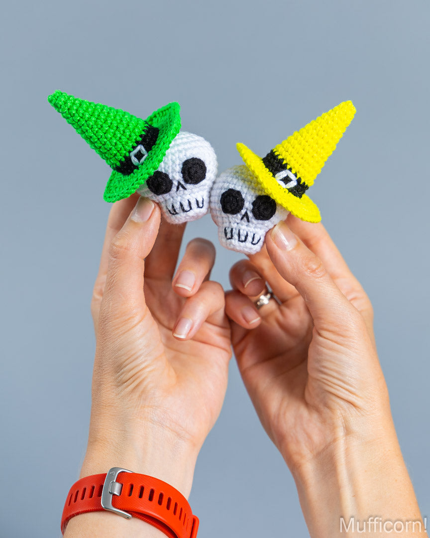 Two hands holding small crocheted skull-shaped accessories with pointed hats, one green and one yellow, against a plain gray background.