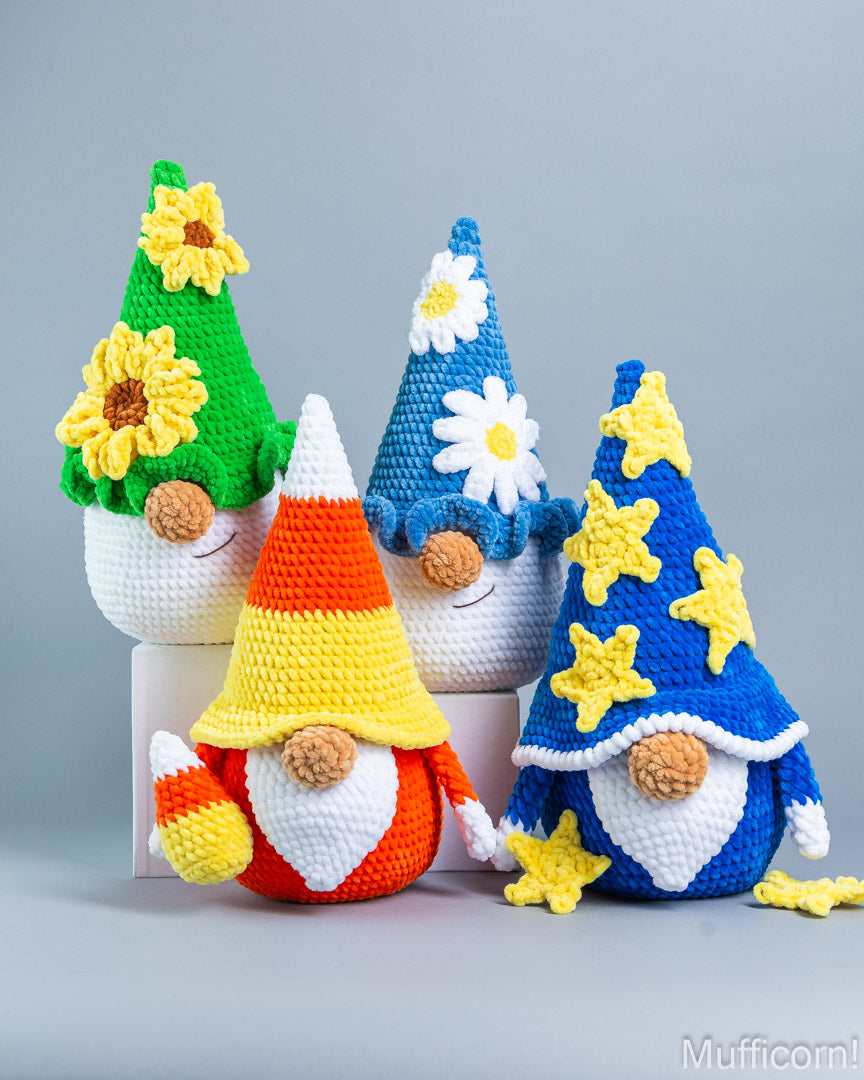 Four crocheted garden gnomes arranged together on a light gray studio background. The gnomes have white beards and colorful pointed hats: one green hat with yellow sunflowers, one blue hat with white daisies, one orange-yellow-white striped cone hat, and one blue hat with yellow stars.