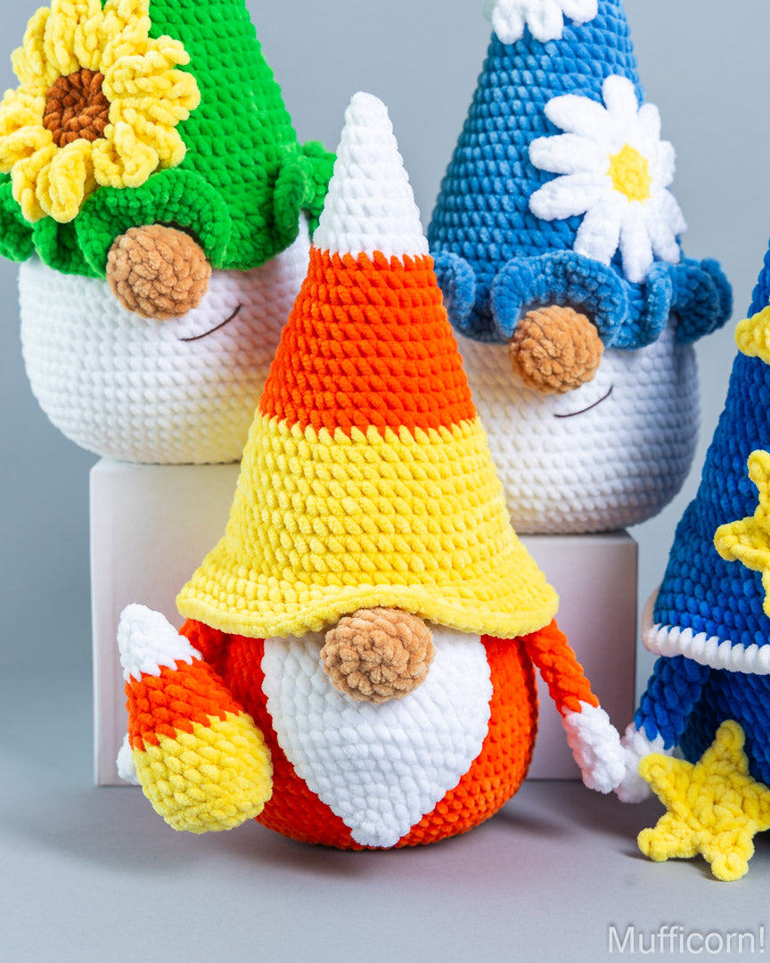 Three crocheted gnome figures with colorful hats and flower details, arranged on white pedestals against a light gray background. The central gnome wears a candy corn hat in orange, yellow, and white, with a white beard and brown nose.
