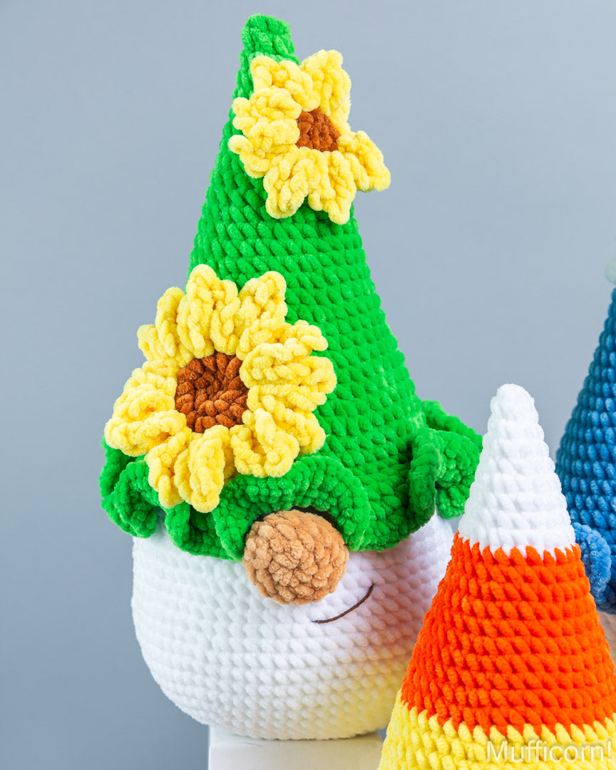 A crocheted gnome figurine with a bright green pointed hat decorated with two yellow sunflowers, a white textured body, and a brown yarn nose, shown against a soft gray background with other colorful crocheted figures partially visible.