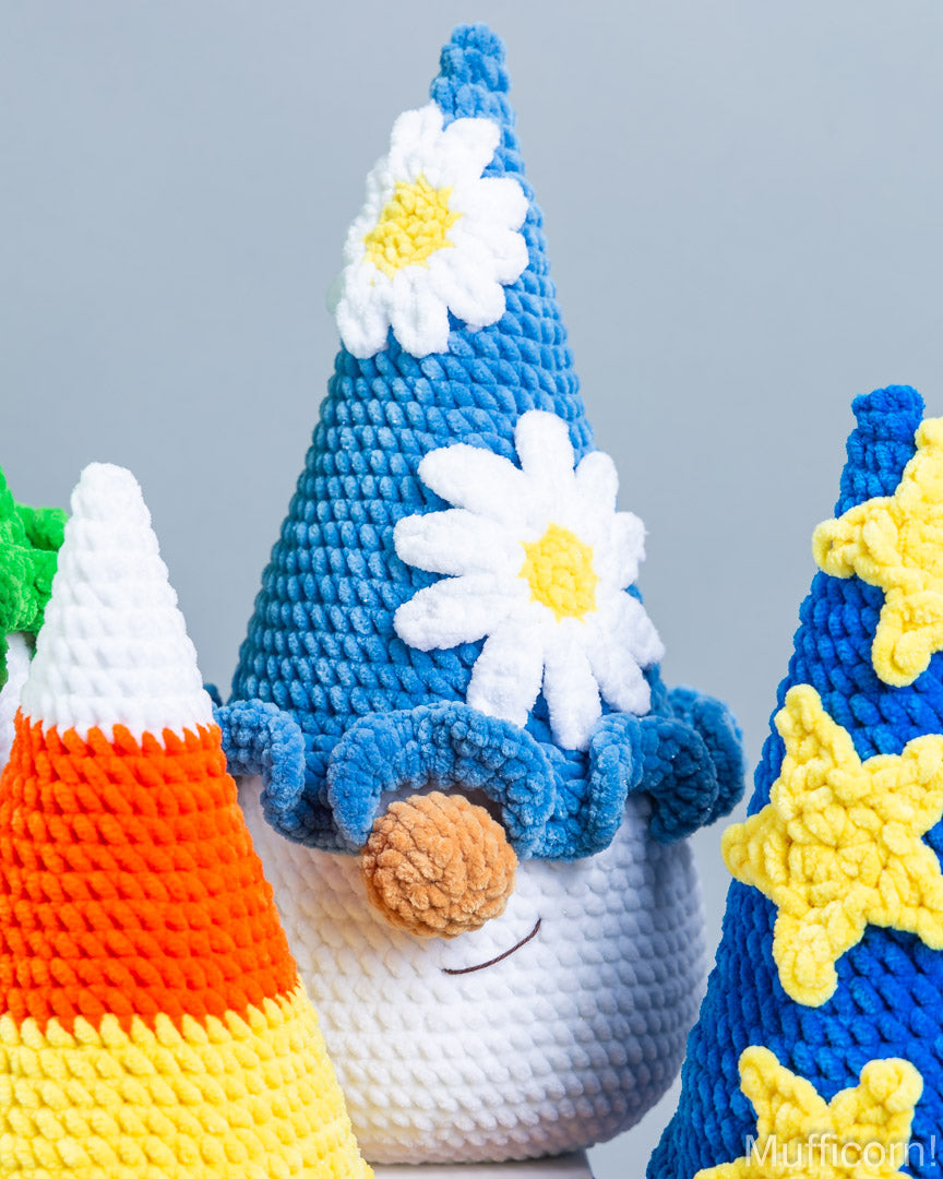 A close-up of crocheted holiday gnome figures, with the central blue hat and white daisy details most prominent against a soft gray background.