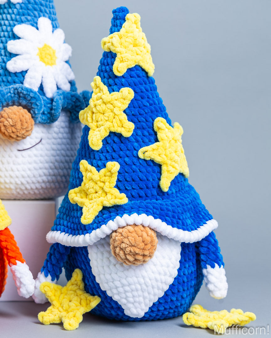 A crocheted gnome doll with a tall blue hat decorated with yellow stars, a white beard, and a pinecone nose, shown on a light gray background with other crocheted figures partially visible.