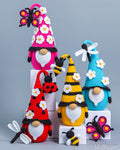 Four crocheted Easter gnomes with colorful pointed hats decorated with flowers, shown on white display blocks against a gray background. A pink gnome with a butterfly, a red gnome with a ladybug and dragonfly, a yellow gnome with a bee, and a blue gnome with a dragonfly are displayed.