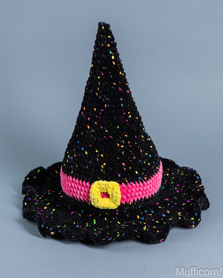 Handmade black knit witch hat with multicolor confetti yarn, a bright pink band, and a yellow crocheted buckle detail, set against a solid gray background.