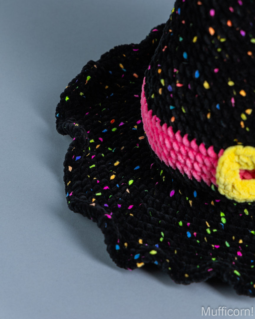 Close-up of a black crocheted hat with multicolored sprinkle details, a pink band, and a yellow pom-pom on a gray background.