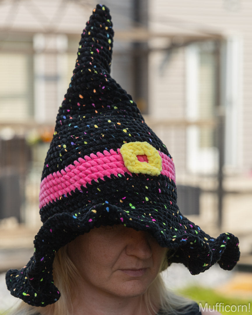 A person wearing a crocheted black witch hat with a bright pink band, a yellow buckle detail, and multicolored confetti accents, photographed outdoors with a blurred background.