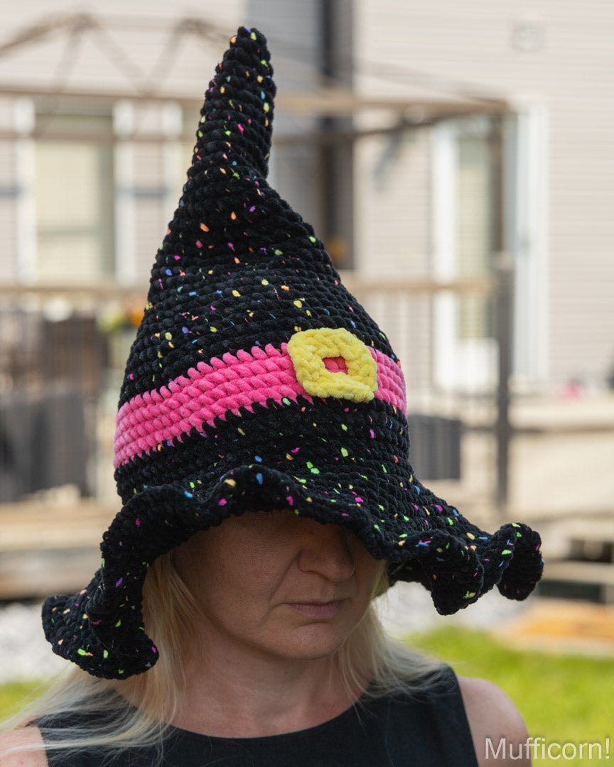 A person wearing a crocheted witch hat in black with a bright pink band, a yellow buckle detail, and multicolored yarn sprinkles, photographed outdoors with a blurred house and patio in the background.