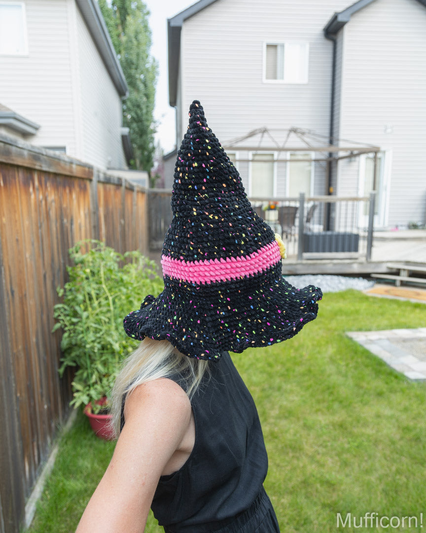 A person standing in a backyard wearing a crocheted witch hat. The hat is black with multicolored yarn speckles and a bright pink band around the middle.